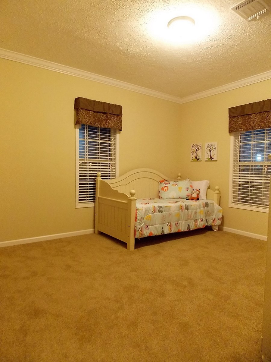 A cozy, warmly lit bedroom with beige walls and carpet. It features a single bed with a colorful, patterned bedspread, pillows, and wall art above.