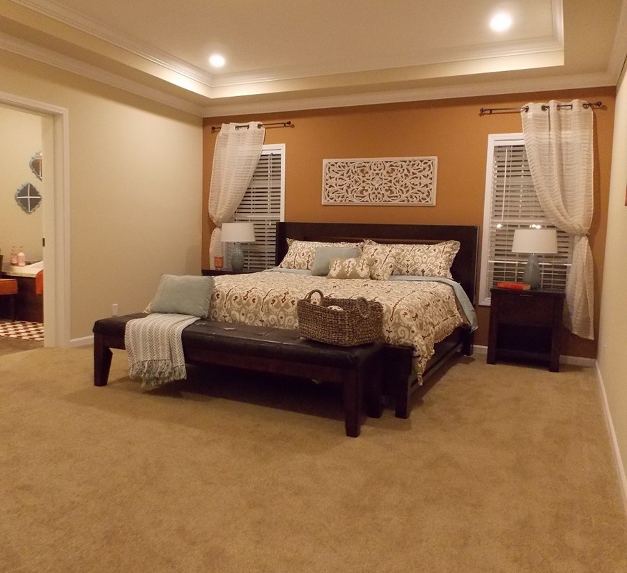 Cozy bedroom with a large bed featuring patterned bedding, flanked by two lamps on nightstands. Beige walls, carpet, and soft lighting create a warm ambiance.
