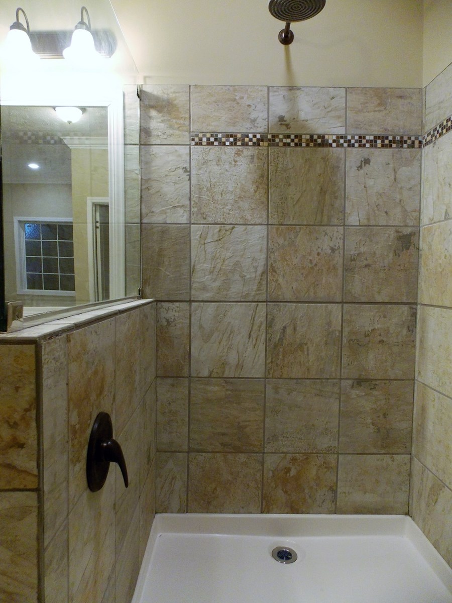 A modern bathroom shower with beige stone tiles, a dark bronze showerhead, and matching faucet. A glass partition and ambient lighting create a serene atmosphere.