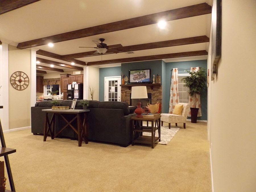 Spacious living room with beige carpet, dark sectional sofa, and rustic wood beams on the ceiling. A stone fireplace, TV, and cozy decoration enhance warmth.