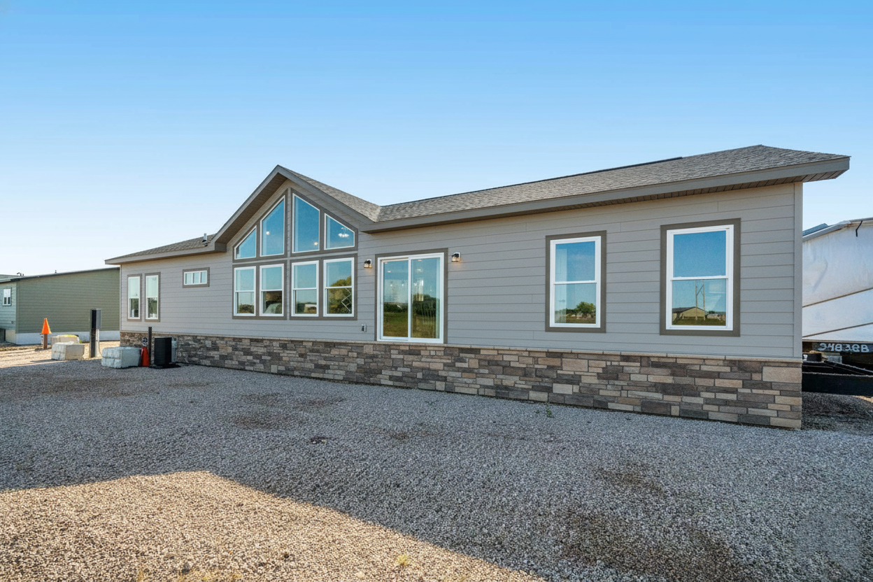 Modern, single-story modular home with gray siding, stone accents, large windows, and a gable roof, set on a gravel lot under a clear blue sky.