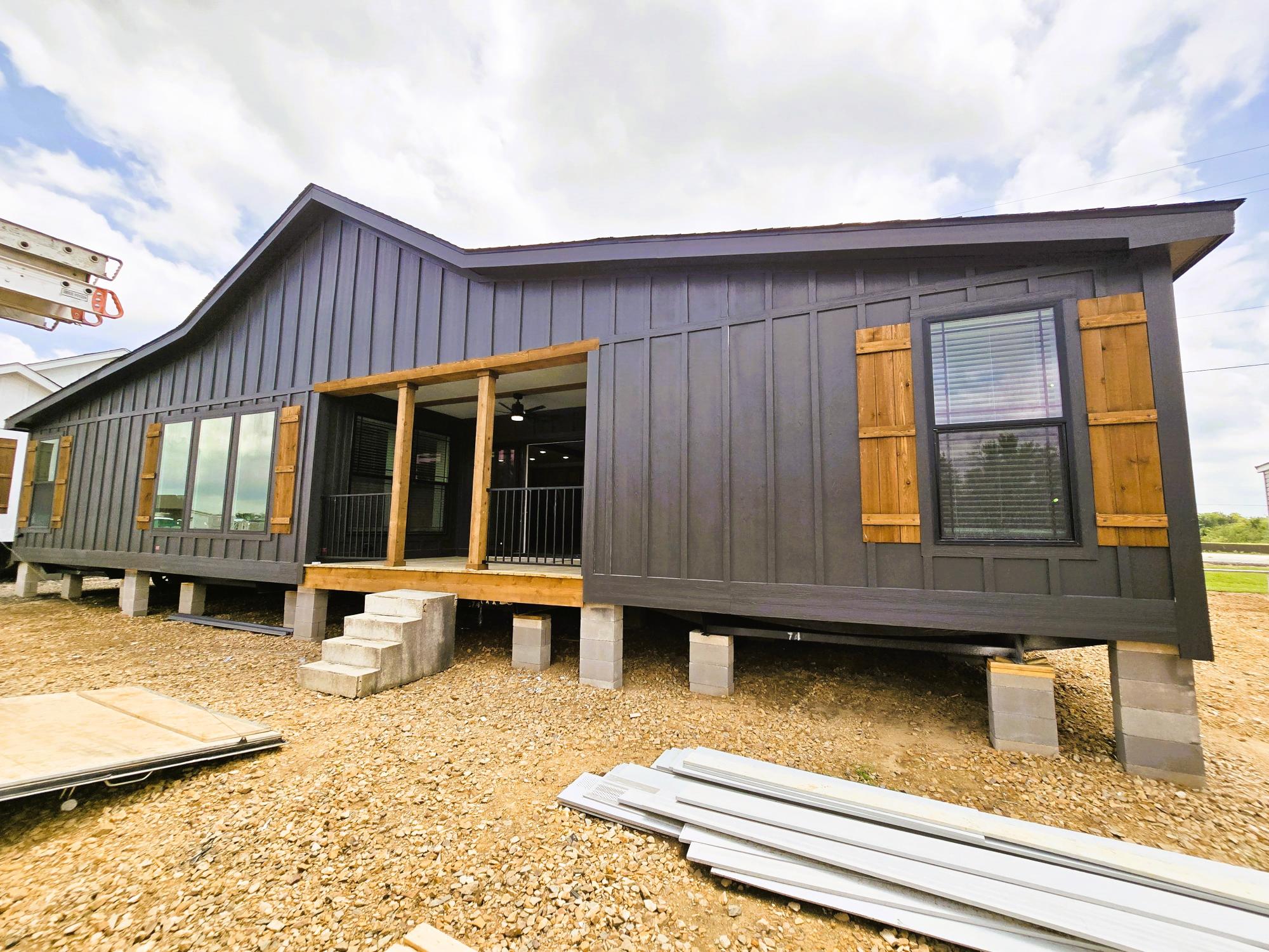 Modern modular home on concrete blocks. Dark wood panels with wooden shutters under a cloudy sky, set on a gravel surface with metal sheets nearby.