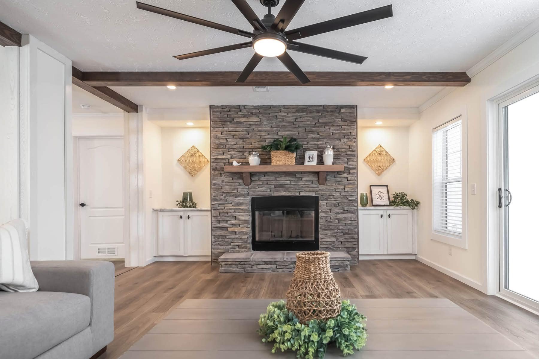 Stylish living room with a stone fireplace, wooden mantel, and built-in cabinets. Features a ceiling fan, light wood floors, and a cozy, modern vibe.