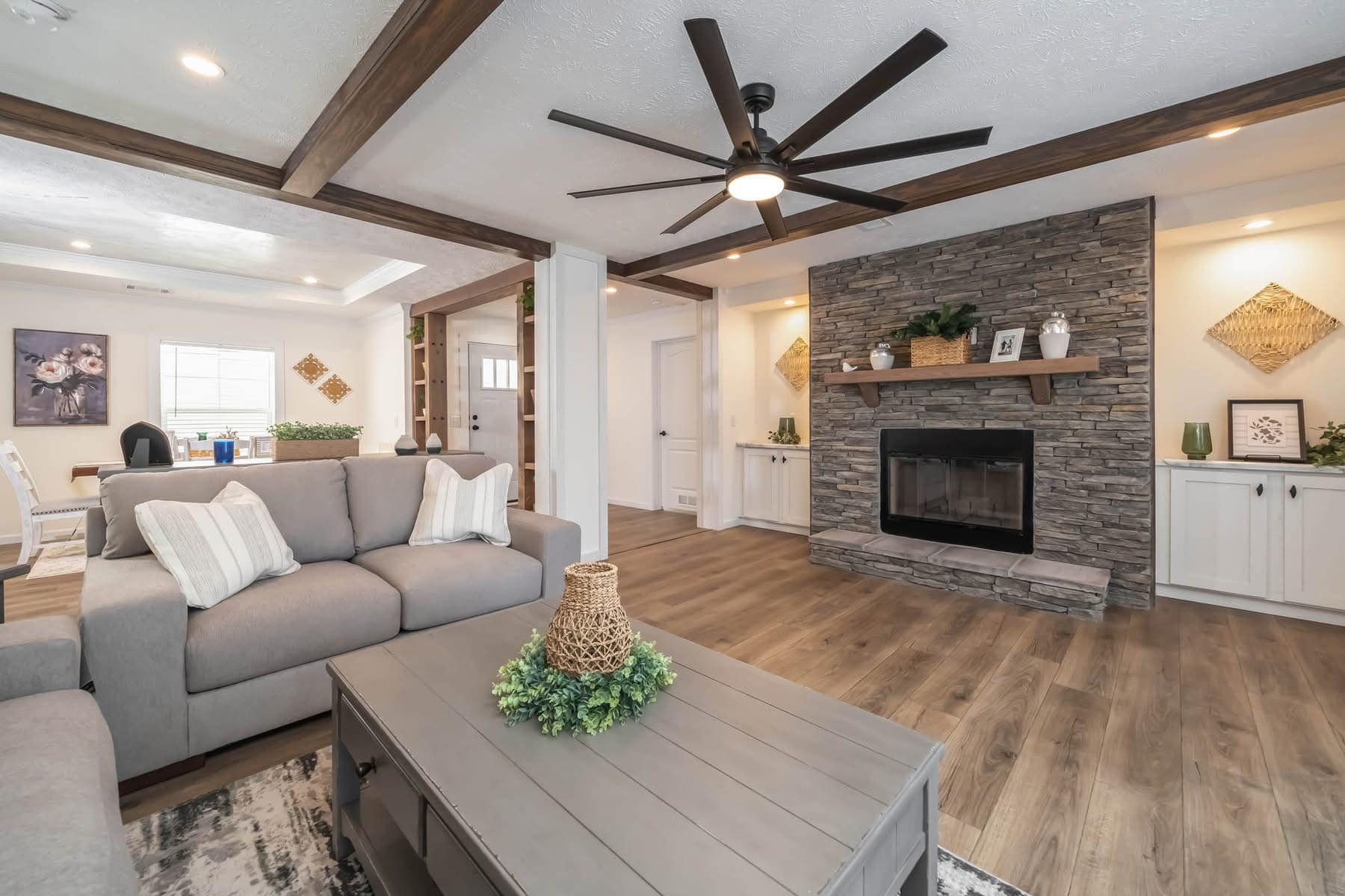 Cozy living room with gray sofas, a stone fireplace, and wood accents. Neutral tones create a calm atmosphere. Ceiling fan and potted plants present.