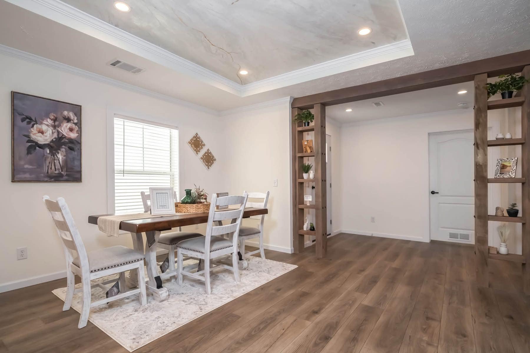 Dining room with a wooden table and six chairs on a textured rug. Soft lighting and pastel walls create a cozy, elegant atmosphere. Artwork features floral designs.