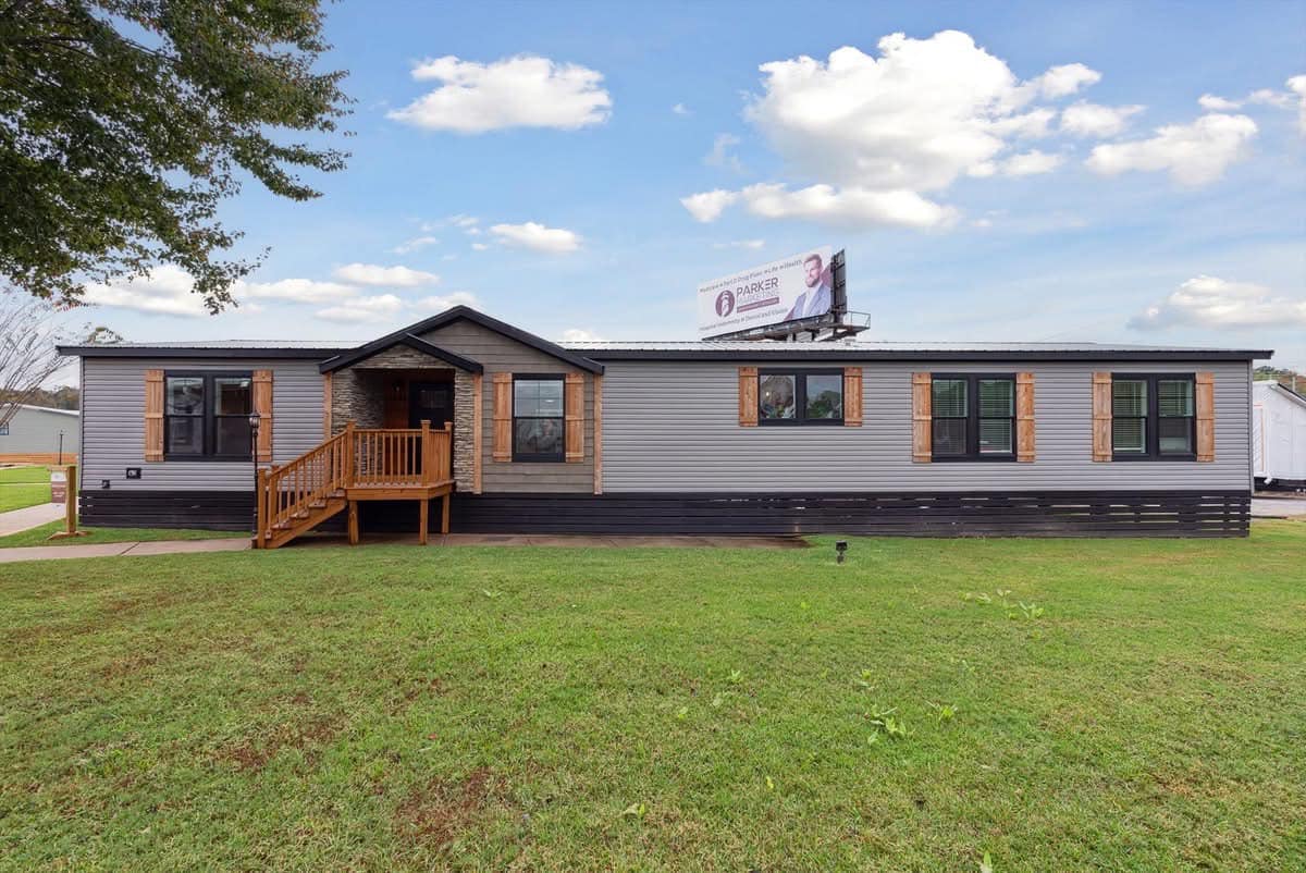 A modern, single-story manufactured home with a gray facade and wooden accents sits on a green lawn. A billboard is visible against a partly cloudy sky.