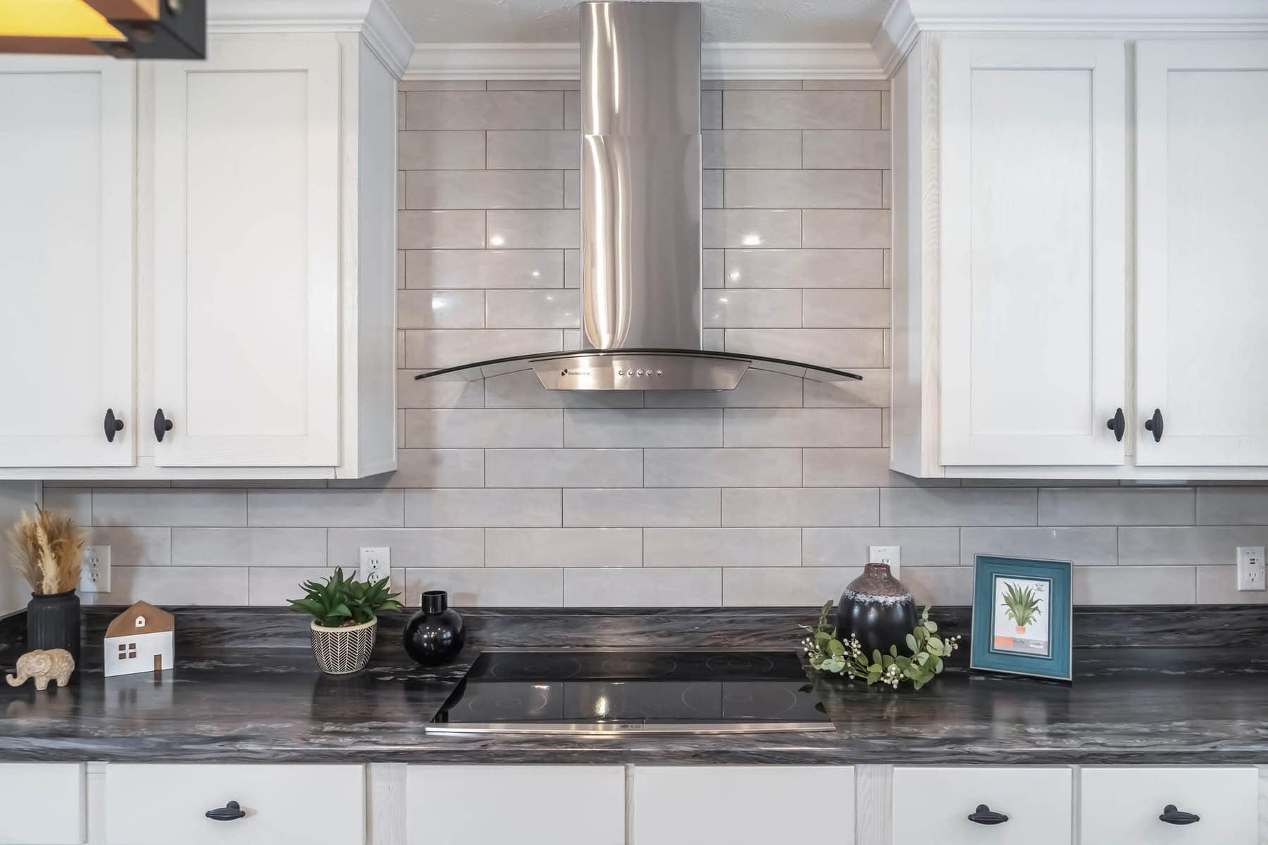 Stylish kitchen with white cabinets, gray tile backsplash, sleek stainless steel hood over a stovetop. Decor includes plants and decorative items.