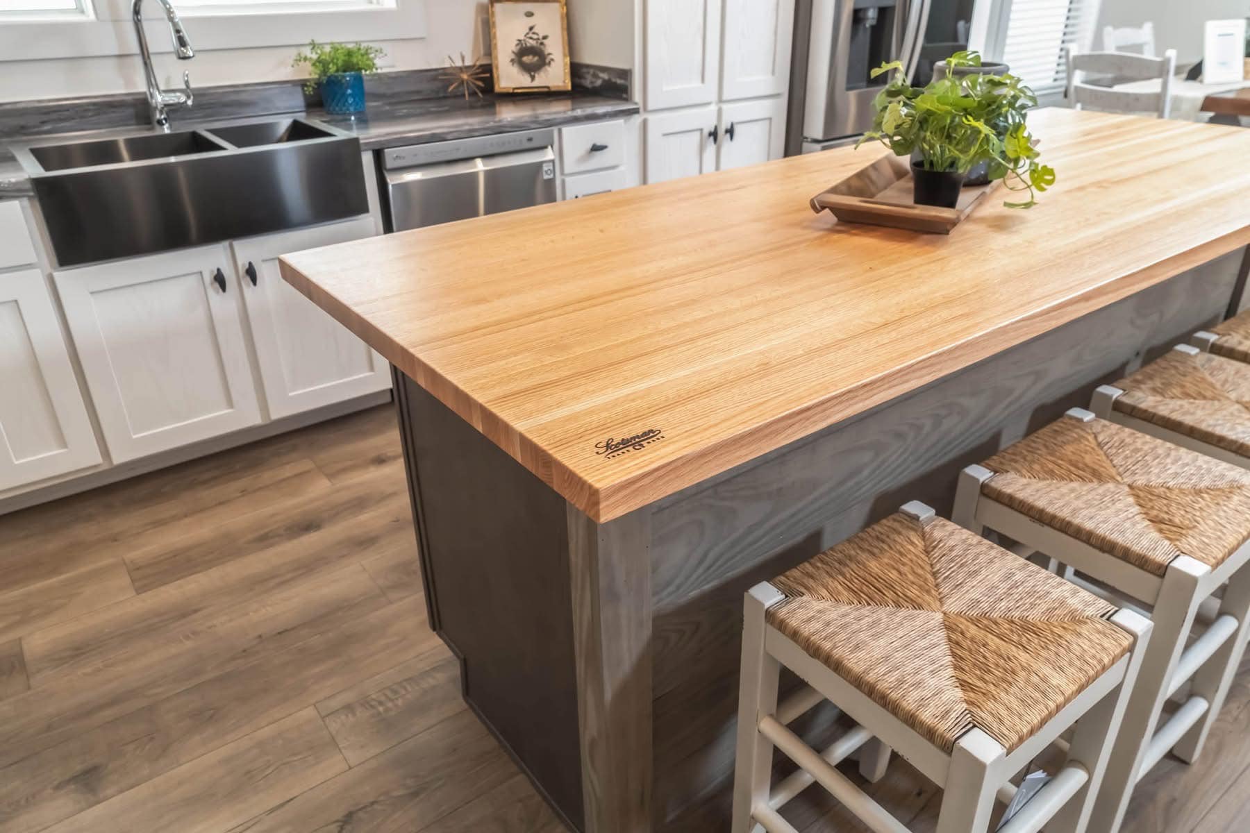 A modern kitchen features a large wooden island with four woven stools, a potted plant centerpiece, white cabinetry, and a farmhouse sink.