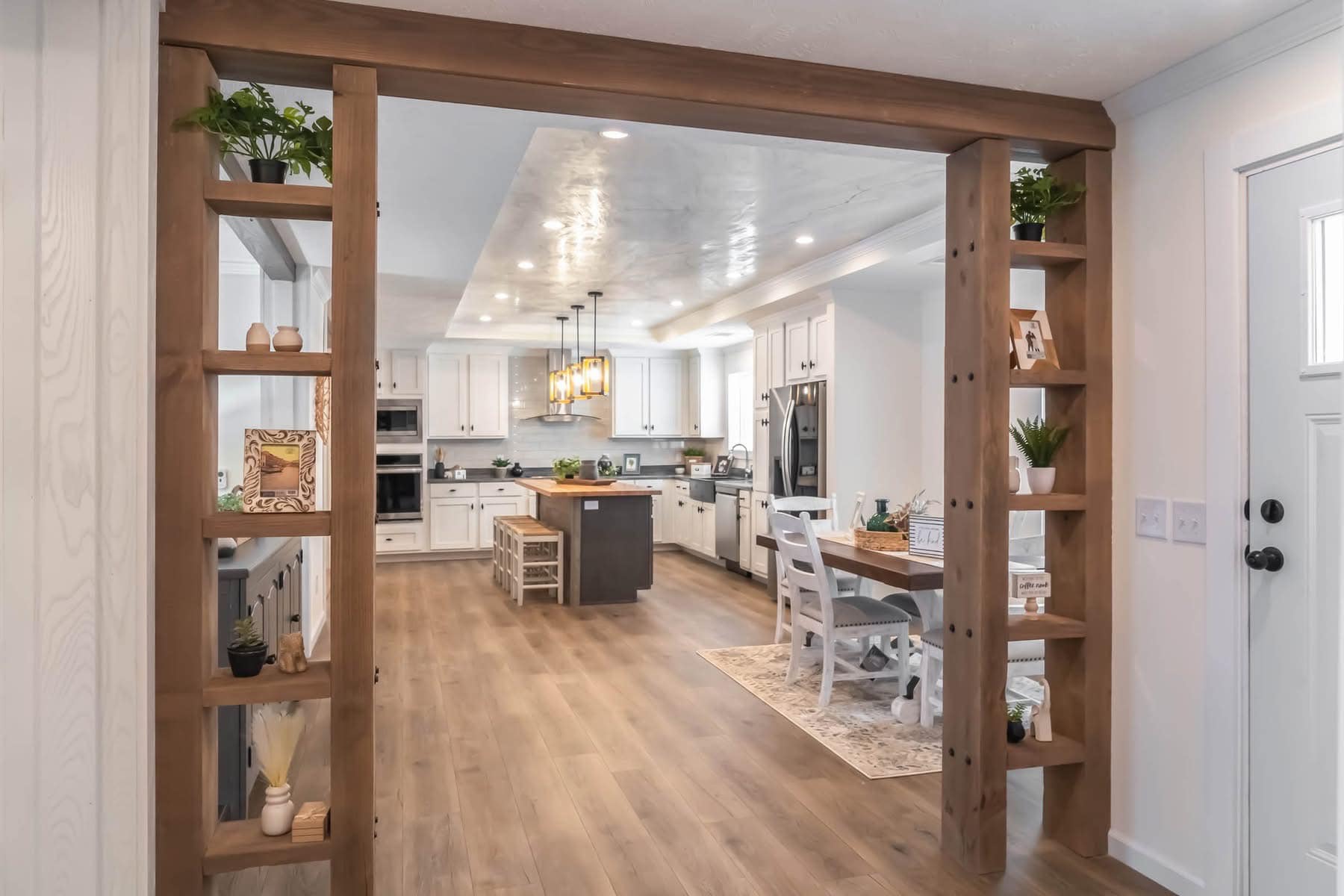 Modern kitchen with wood flooring, white cabinets, and a central island with stools. Open shelving holds plants and decor. Bright lighting adds warmth.