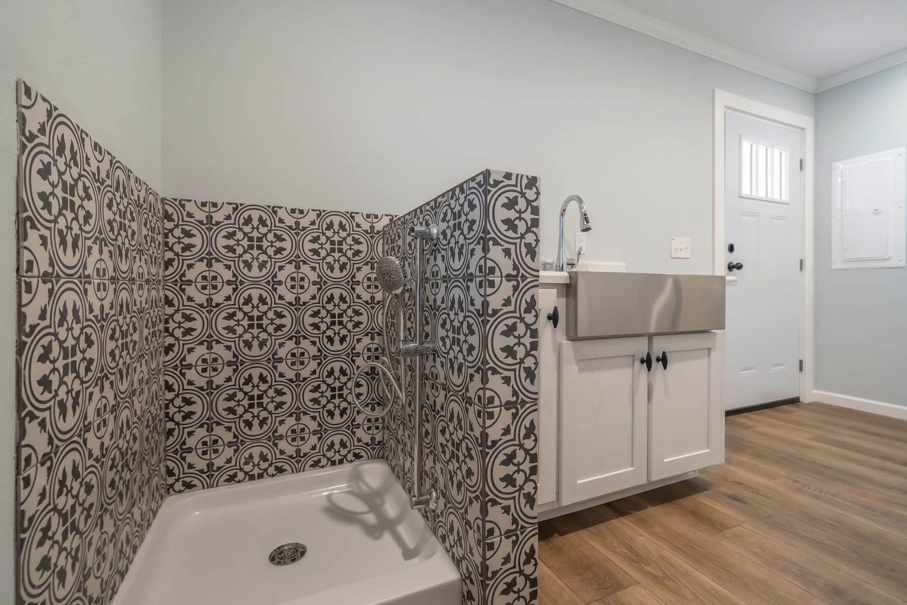 Tiled shower with intricate black and white design next to a modern utility sink in a light blue room with wooden floors and a white door.