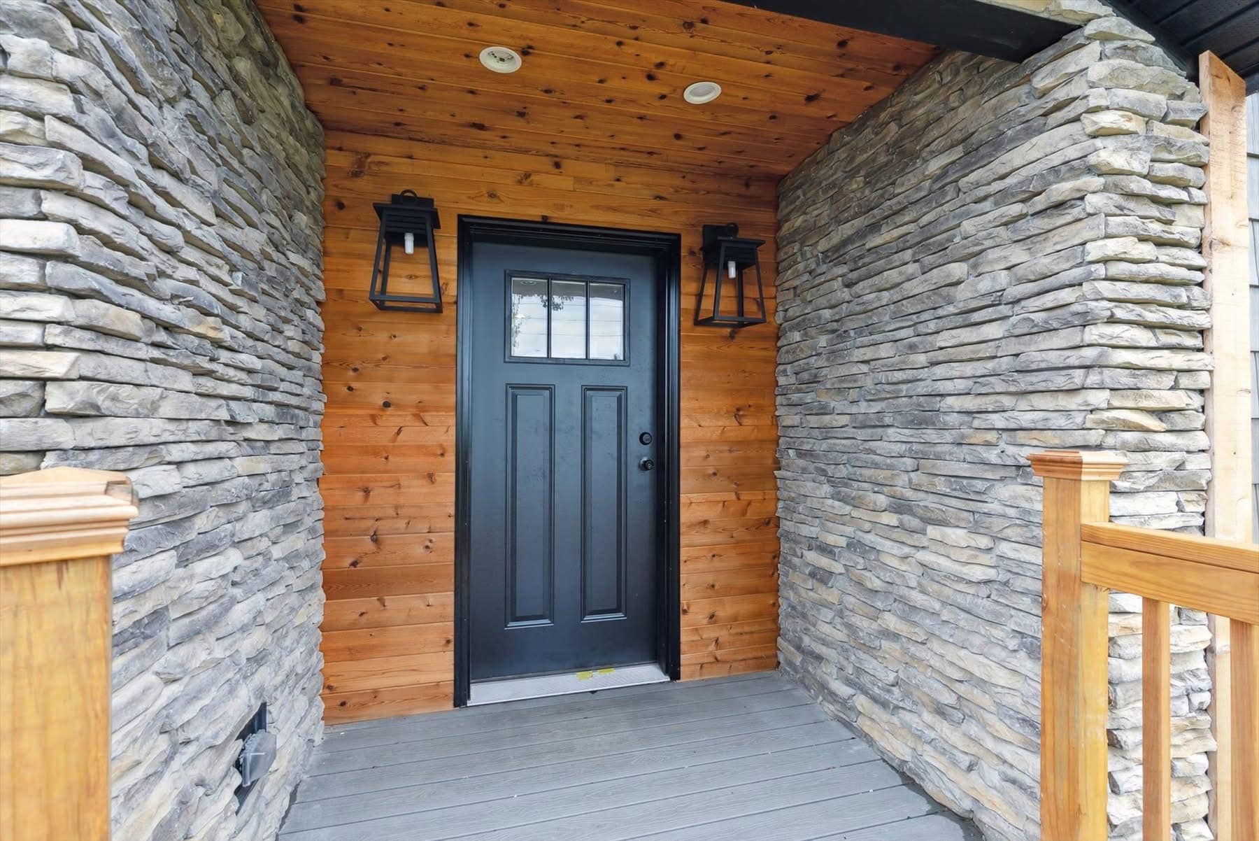Stone porch with a wooden ceiling and walls, featuring a black door flanked by lantern-style lights, conveys a rustic and welcoming atmosphere.
