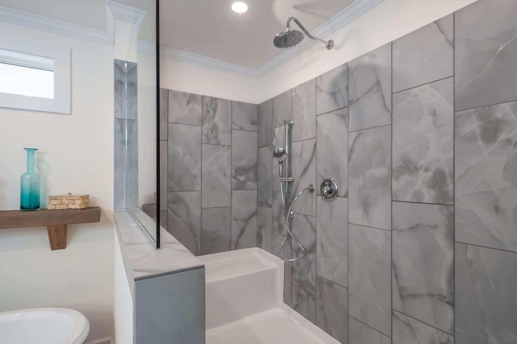 Modern bathroom with gray marbled tiles, a chrome showerhead, and a built-in white bench. Wooden shelf holds blue glass vase and wicker box.
