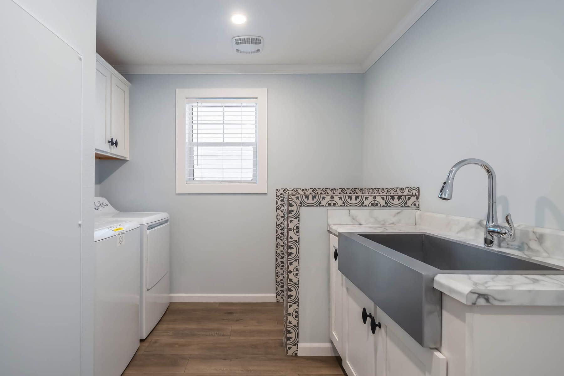 Bright laundry room with a white washer and dryer on the left, a large stainless steel sink with marble countertop on the right, and patterned tile accent.