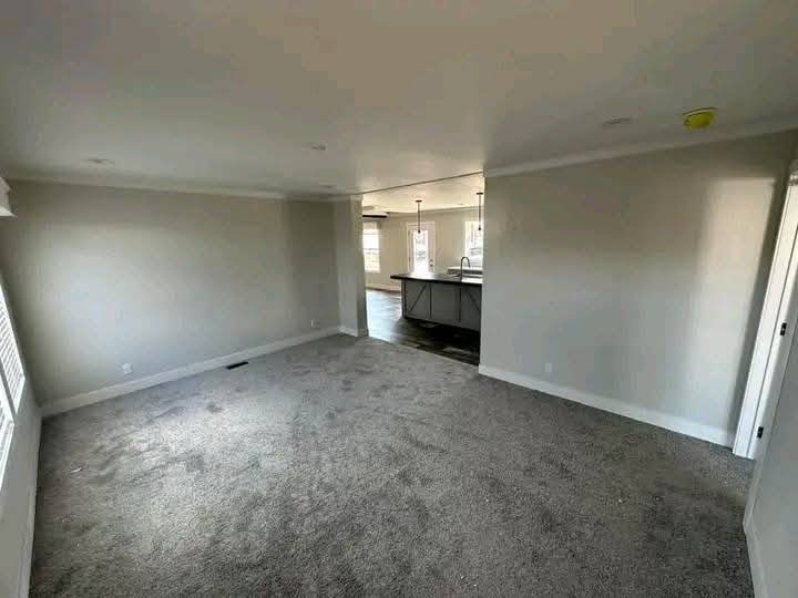 Spacious room with gray carpet, light gray walls, and abundant natural light. Kitchen with pendant lights visible in the background, creating a cozy atmosphere.