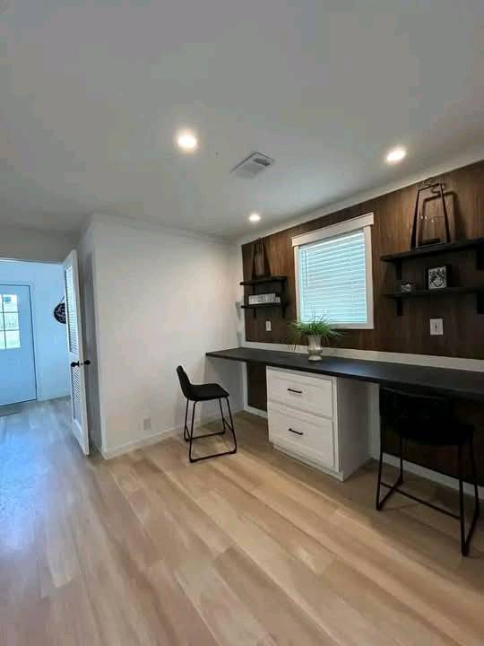 Modern office with wooden flooring, a long desk, and two black chairs. Shelves adorn a dark accent wall with a window, creating a clean, cozy atmosphere.