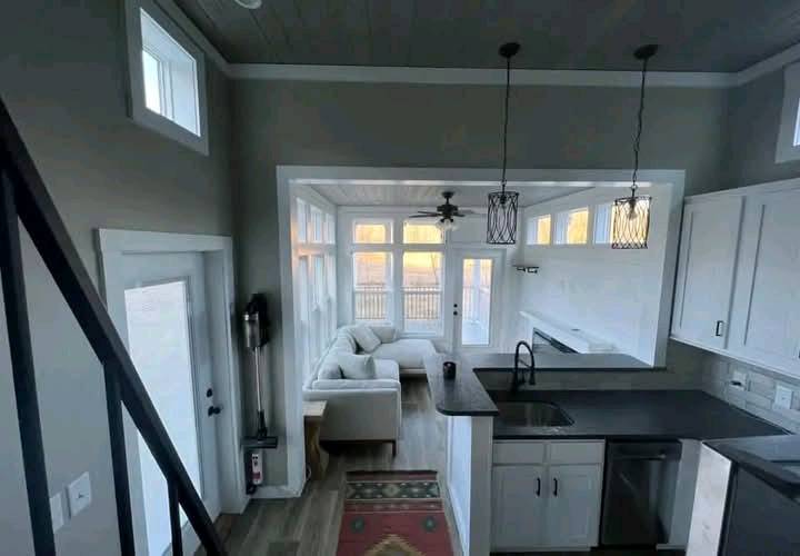 Modern living space with high ceilings, featuring a cozy white sofa in a sunlit room with large windows. A sleek kitchen with black countertops is visible.