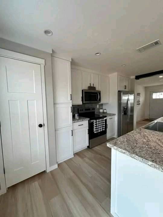Modern kitchen with light wood flooring, white cabinets, granite countertops, and stainless steel appliances. A door is on the left, creating an open, airy feel.