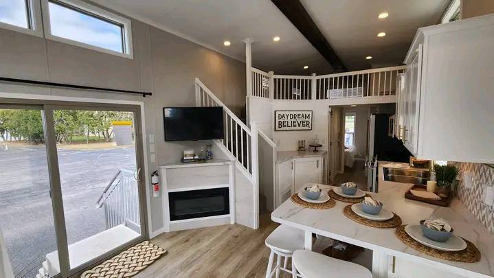 Modern tiny home interior with open layout. Features light wood flooring, white kitchen cabinets, breakfast bar with stools, and loft with "Daydream Believer" sign. Large windows provide natural light and outdoor view. Cozy, inviting atmosphere.