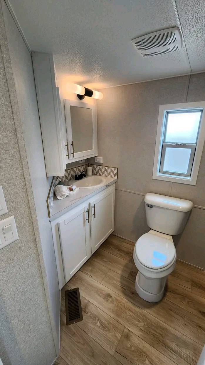Small modern bathroom with a white toilet, white vanity with sink, and mirror cabinet. Light wood flooring and soft lighting create a clean, cozy ambiance.