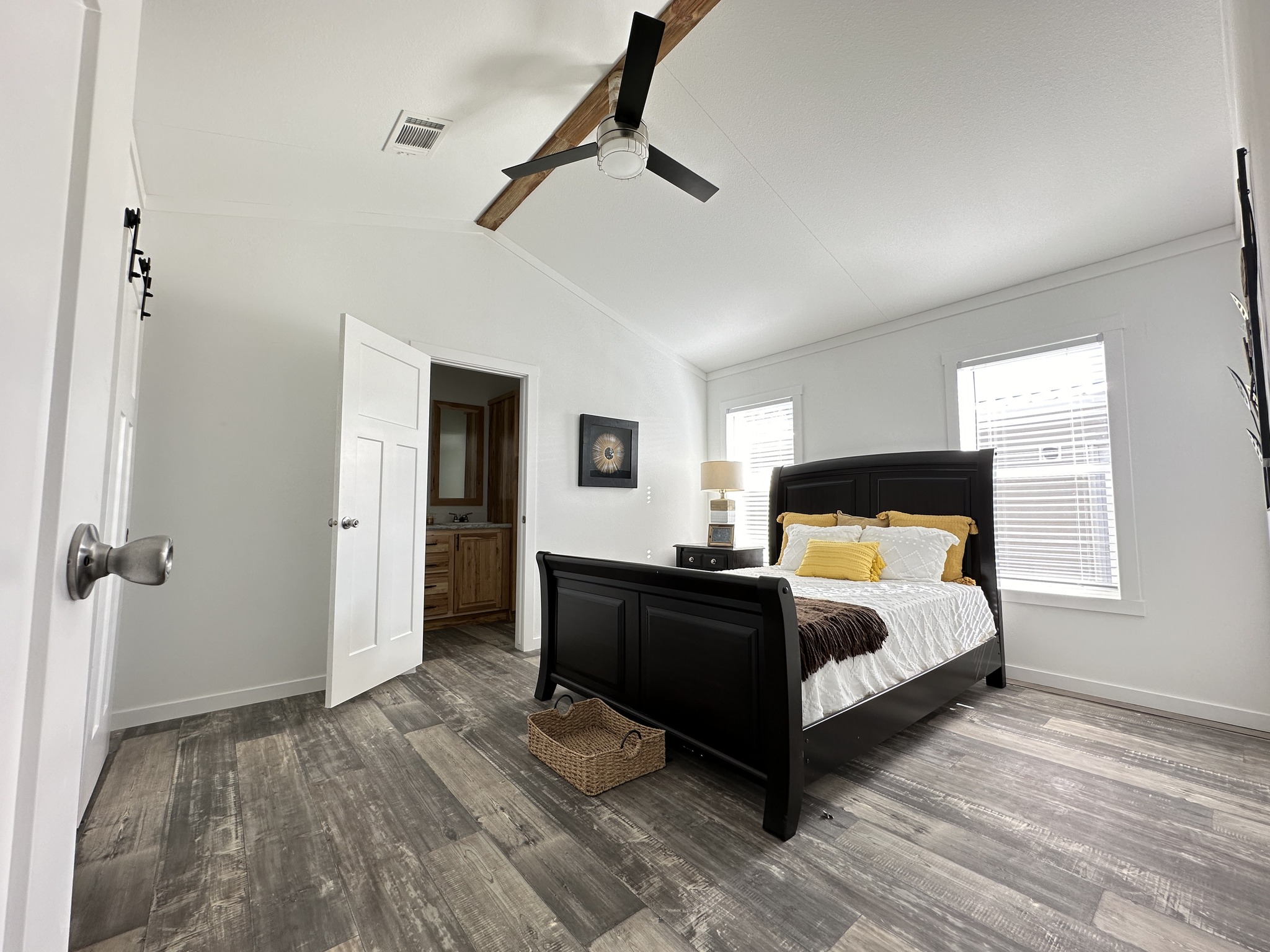 A cozy bedroom with a black wooden bed, white and yellow bedding, and a brown throw. Two windows, a ceiling fan, and a wicker basket enhance the serene ambiance.
