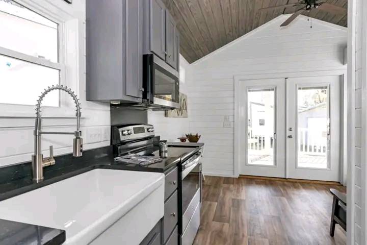 Modern kitchen with black countertops, farmhouse sink, and stainless steel appliances under gray cabinets. A wooden ceiling adds warmth. French doors lead outside, letting in natural light.
