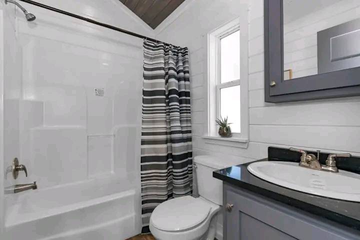 A small bathroom with a white bathtub and striped shower curtain. There's a toilet under a window, and a gray vanity with a black countertop enhances the modern, clean design.