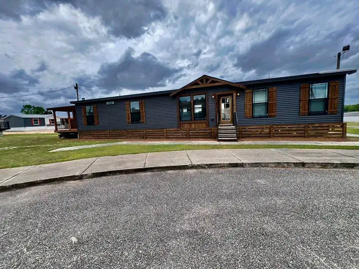 A dark gray modular home with a wooden porch and shutters stands against a cloudy sky. The scene is calm, with a curved sidewalk and grassy lawn.