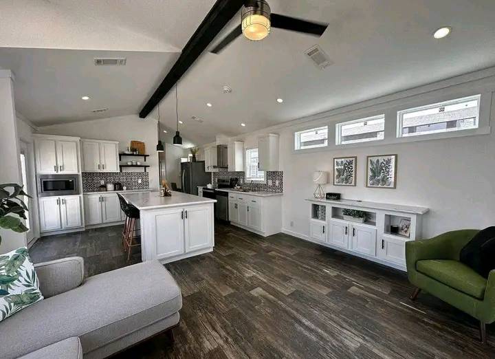 Modern open-plan kitchen and living area with white cabinets, black pendant lights, and wood flooring. Natural light streams through small windows, creating an inviting ambiance.