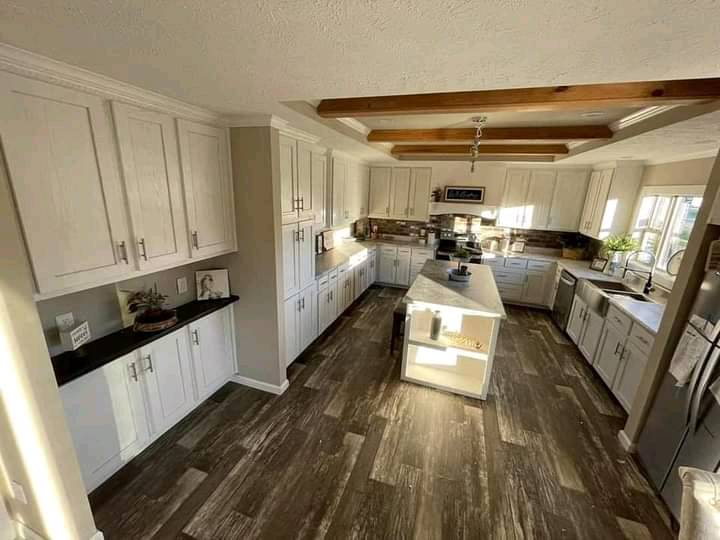 Spacious kitchen with white cabinets, an island with hanging lights, and wooden beams on the ceiling. Sunlight creates a warm, inviting ambiance.