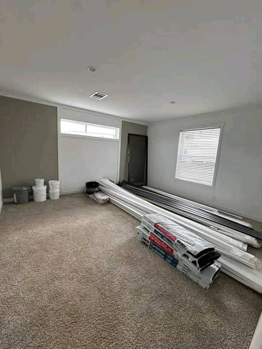 A spacious, empty room with beige carpet and white walls, containing stacked PVC pipes and buckets in a corner. Large windows let in natural light.