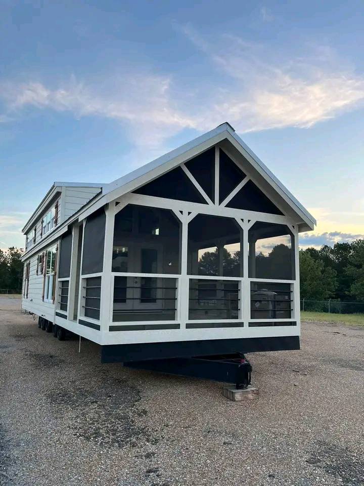 A tiny house on wheels with a white frame and screened porch sits on a gravel lot. The sky is clear, suggesting a peaceful, serene evening atmosphere.