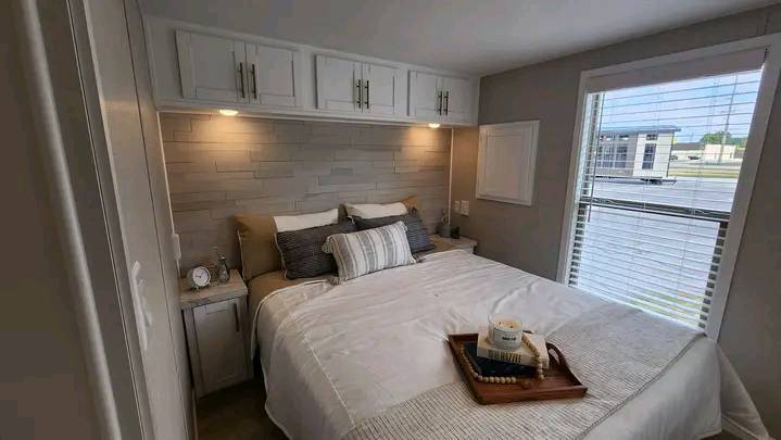 A cozy bedroom with a neatly made bed, white linens, and decorative pillows. Warm lighting from above cabinets creates a soft, inviting atmosphere.