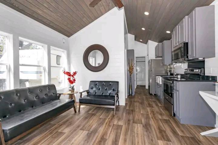 Modern living room and kitchen with dark wood flooring, gray cabinetry, black sofas, circular mirror, red flowers, and natural light from large windows.