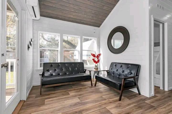 Inviting sunroom with leather sofas, large windows, and wood floors. A round mirror and bright red flowers on a glass table add charm.