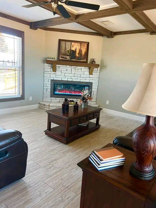 Cozy living room with a stone fireplace and modern electric insert, wooden beams on the ceiling, and elegant dark wood furniture with decorative items.