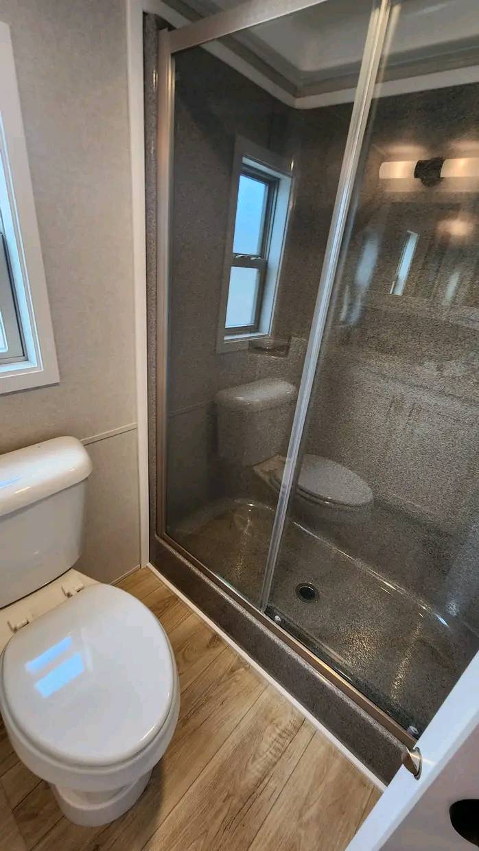 A modern bathroom featuring a white toilet, wood-patterned floor, and a glass-enclosed shower. The room feels clean and minimalistic, with neutral tones.