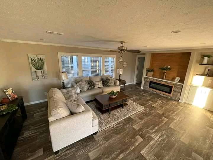 Cozy living room with beige walls, a light sectional sofa, wooden coffee table on a rug, modern fireplace, ceiling fan, and decorative plants.