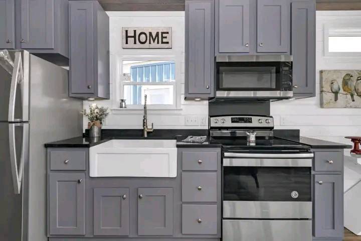 Modern kitchen with gray cabinets, black countertop, and stainless steel appliances. A "Home" sign above the window adds a cozy touch.