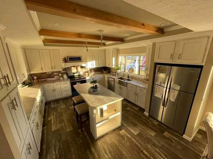Spacious kitchen with warm lighting, featuring white cabinets, a central island with stools, stainless steel appliances, and wooden ceiling beams.