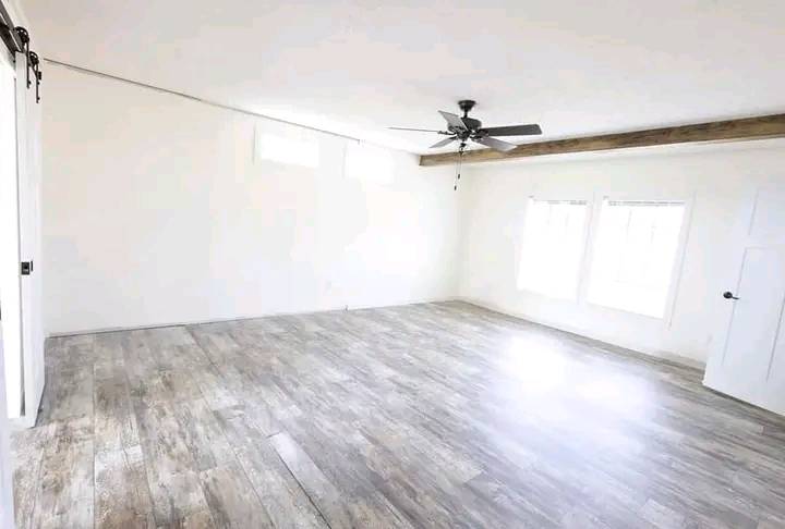 Spacious, empty room with light wood flooring, white walls, two large windows, a ceiling fan, and a wooden ceiling beam, creating an airy, minimalist feel.