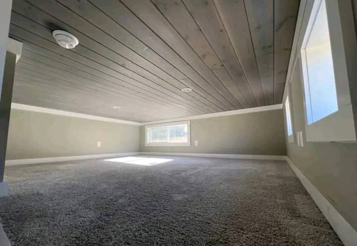 Cozy attic space with low wooden ceiling, carpeted floor, and small windows on each side letting in soft natural light, creating a serene atmosphere.