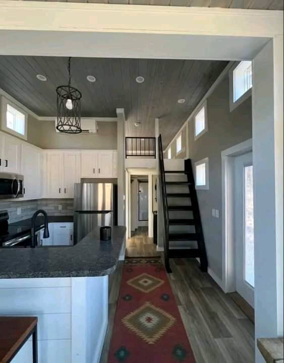 A cozy kitchen with white cabinets and a gray countertop leads to a wooden staircase. Light from small windows creates a warm, inviting atmosphere.