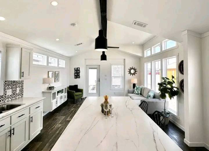 Modern white kitchen and living area with a marble island, pendant light, and large windows. Decor includes a green armchair and indoor plant.