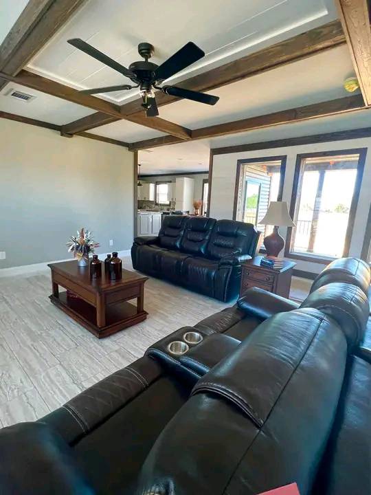 Spacious living room with black leather sofas and a wooden coffee table on a light tile floor. Ceiling fan above, large windows, cozy and inviting.