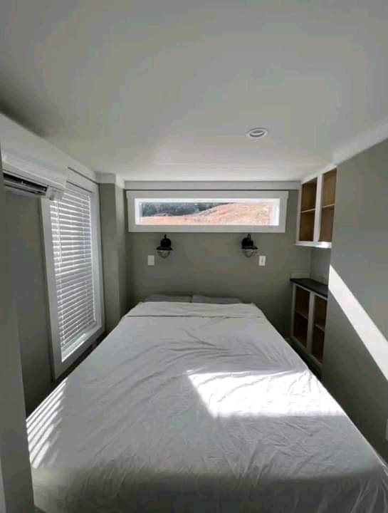 Small, cozy bedroom with a neatly made bed, white linens, and natural light from a thin horizontal window. Shelves and modern lamps add simplicity.