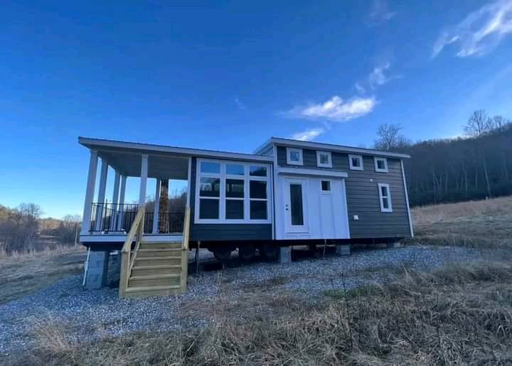 A modern tiny house on a raised foundation in a field under a blue sky. It features a front porch, multiple windows, and a neutral facade, conveying a peaceful rural setting.