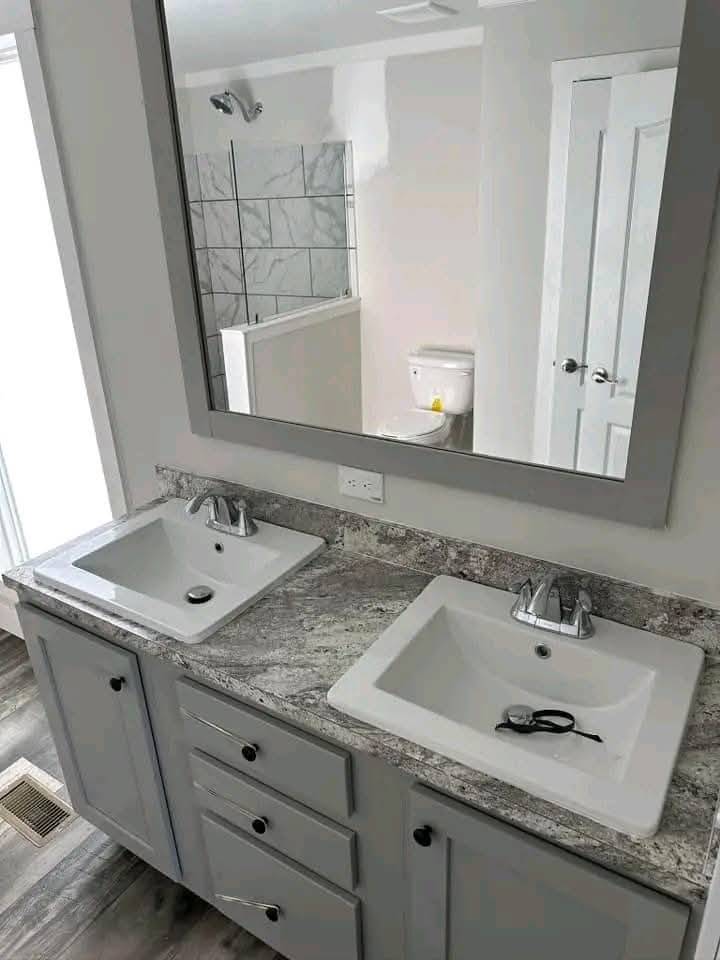 Modern bathroom with a large mirror reflecting a toilet and shower. Features two rectangular sinks on a marble countertop, under bright natural light.