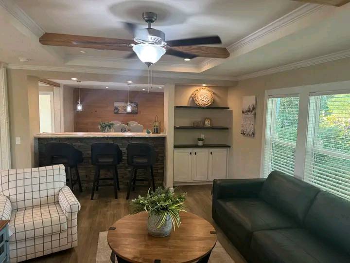 Cozy living room with wooden floor, ceiling fan, plaid armchair, gray sofa, and round coffee table. Kitchen with bar stools in background. Natural light from large windows.
