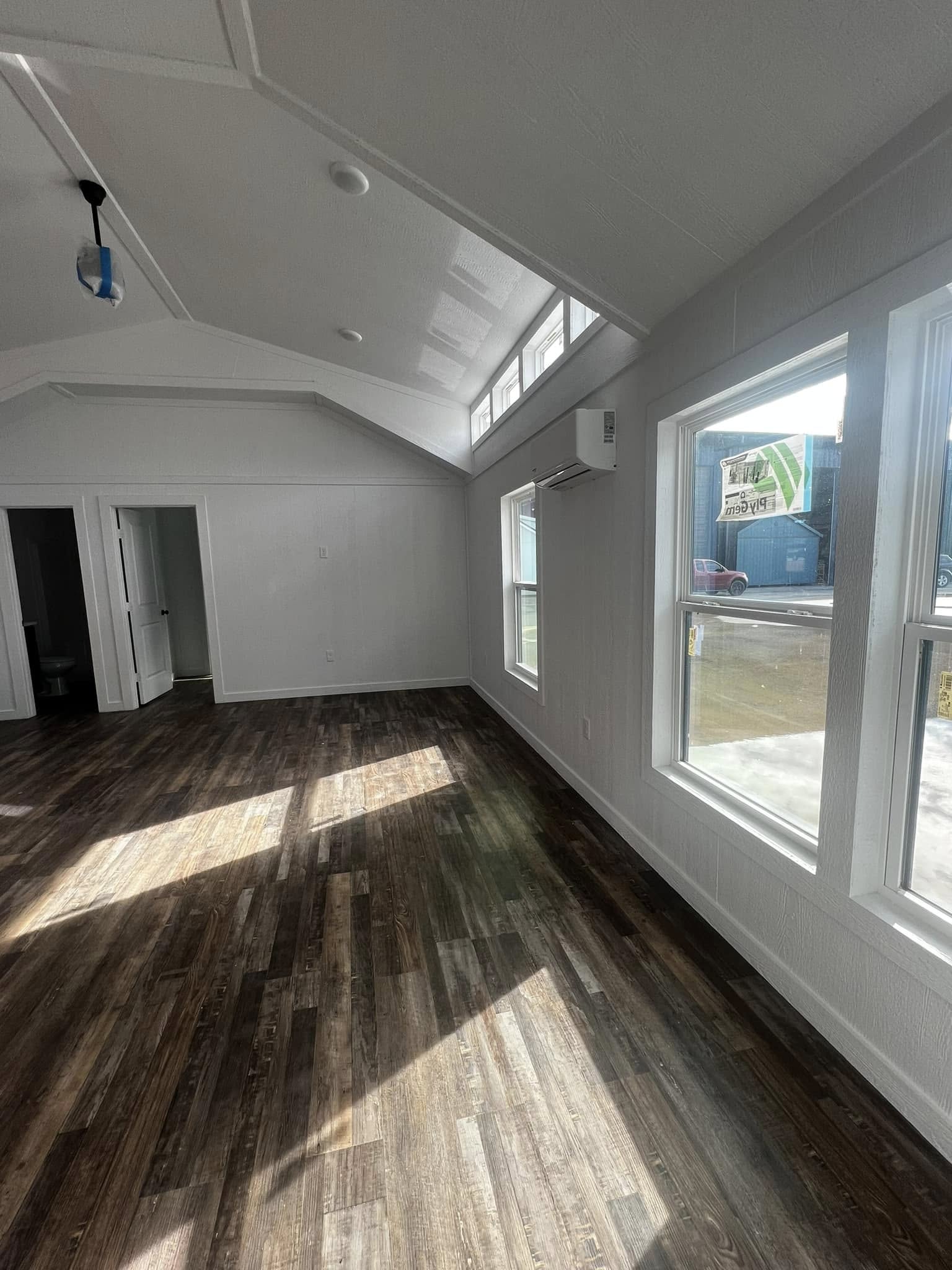Spacious, sunlit room with white walls, high ceiling, and large windows. Dark wood flooring and open doorway leading to another room. Airy and modern.