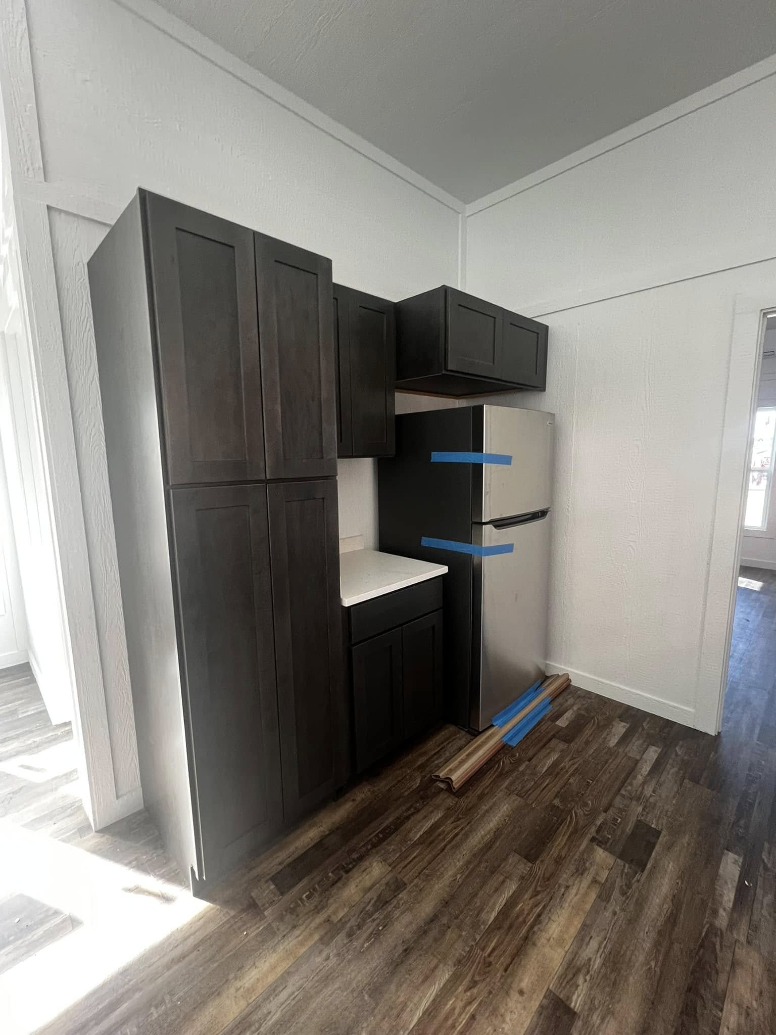 Dark wooden kitchen cabinets and a stainless steel fridge with blue tape are set against white walls and wood-patterned flooring, in a well-lit room.
